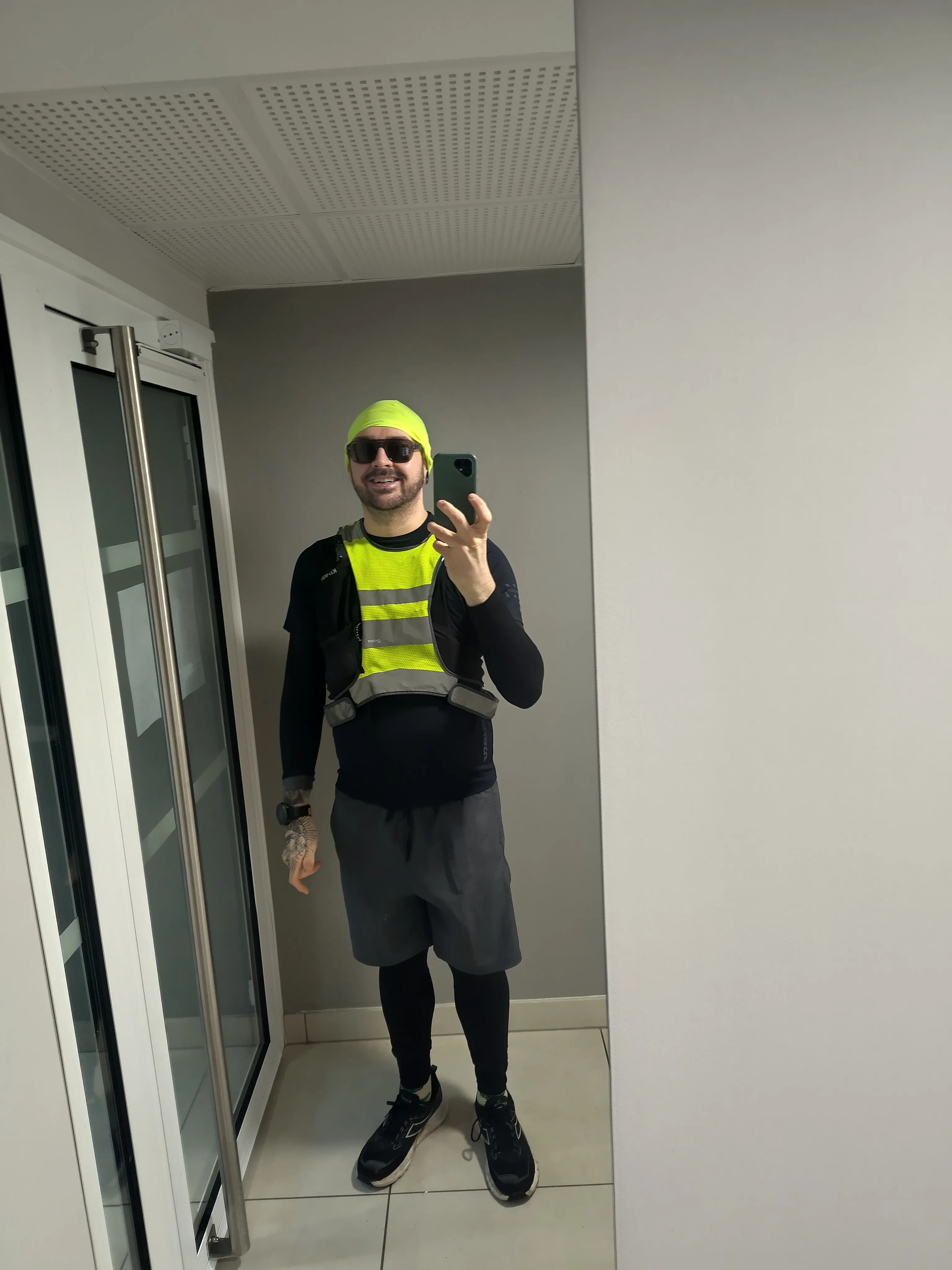 Portrait de moi-même, souriant en tenue de course hivernale noire avec des chaussures noir, devant le miroir dans l'entrée de l'immeuble.